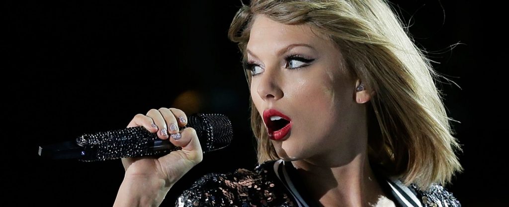 Taylor Swift está cantando. Ela pode ser vista olhando para a esueda com o microfone na mão. A cantora, loira, está usando um batom vermelho.