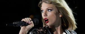 Taylor Swift está cantando. Ela pode ser vista olhando para a esueda com o microfone na mão. A cantora, loira, está usando um batom vermelho.