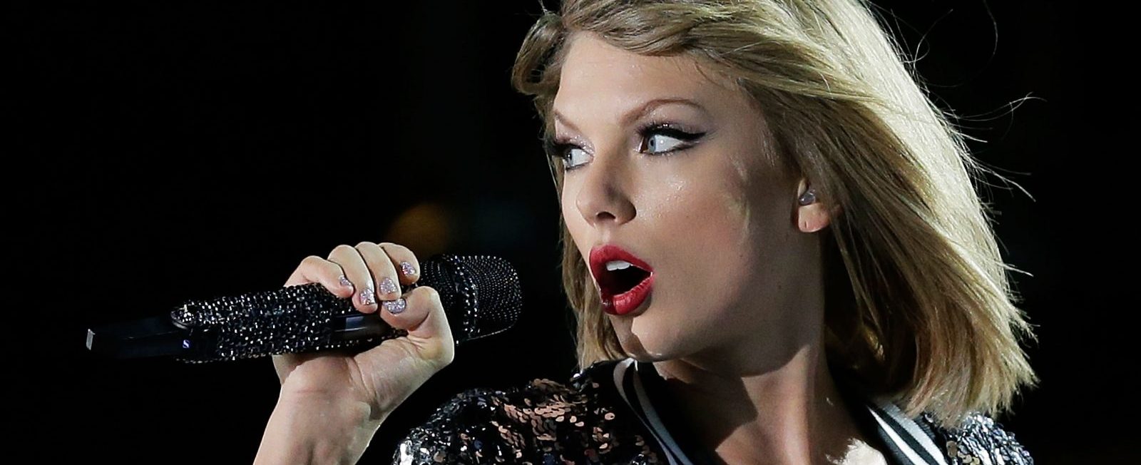 Taylor Swift está cantando. Ela pode ser vista olhando para a esueda com o microfone na mão. A cantora, loira, está usando um batom vermelho.