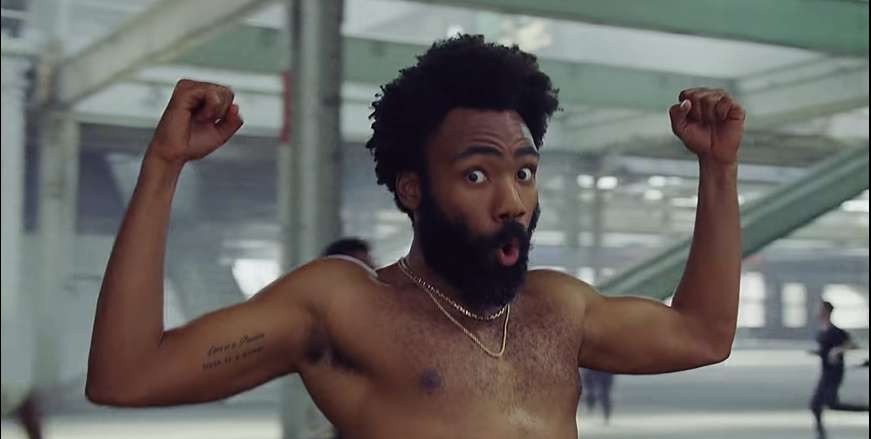 Donald Glover no clipe de This Is America, olhando para a câmera enquanto flexiona os bíceps.