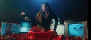 Pitty no clipe de Na Tela, sentada no chão, com uma saia longa vermelha que se espalha no chão e com várias TVs ao fundo, mostrando rostos, bocas e ruídos brancos.