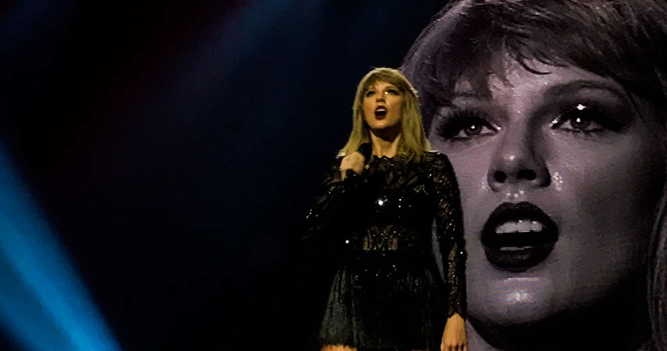 taylor swift no palco da reputation tour olhando para o público suada e com uma roupa toda preta. atrás, há um telão exibindo o rosto da cantora imagem ilustrativa texto sobre taylor swift e a big machine em músicas do folklore