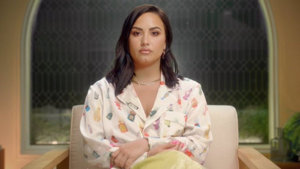 A imagem promocional de "Dancing With The Devil", documentário de Demi Lovato, apresenta a cantora sentada em uma poltrona da cor branca com as pernas cruzadas, enquanto veste uma calça amarela e blusa de mangas compridas colorida. Suas mãos estão sob seu colo e ela olha fixamente para a frente com uma cara séria.