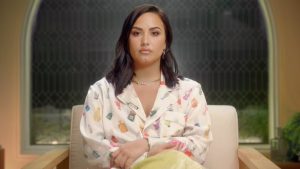 A imagem promocional de "Dancing With The Devil", documentário de Demi Lovato, apresenta a cantora sentada em uma poltrona da cor branca com as pernas cruzadas, enquanto veste uma calça amarela e blusa de mangas compridas colorida. Suas mãos estão sob seu colo e ela olha fixamente para a frente com uma cara séria.
