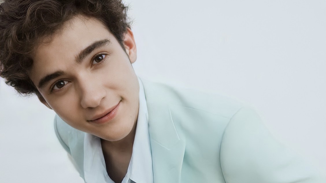 Em um fundo branco, Joshua Bassett aparece de terno azul com uma camiseta branca por dentro. Na imagem, Joshua Bassett está sorrindo para frente enquanto promove seu primeiro EP.