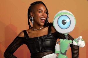 foto de divulgação da karol conká no bbb. a cantora aparece num fundo alaranjado com um boneco do programa ao lado