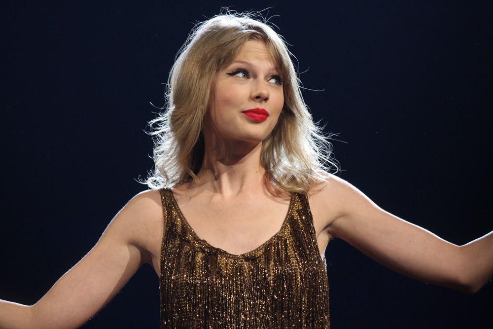 taylor swift sorrindo e de braços abertos em um show de sua turnê imagem ilustrativa do texto sobre as taylors version