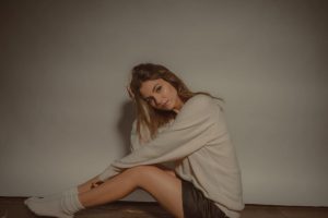 A imagem mostra a atriz Victoria Justice sentada e virada para a esquerda, com os joelhos meio levantados. Ela está com o braço esquerdo apoiado no joelho esquerdo, enquanto sua mão direita está na cabeça. Ela veste um moletom cinza, um shorts preto e uma meia branca. O chão é de madeira e a parede ao fundo tem a mesma cor de sua camisa, cinza. A imagem destaca o post sobre falsas rivalidades femininas.