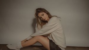 A imagem mostra a atriz Victoria Justice sentada e virada para a esquerda, com os joelhos meio levantados. Ela está com o braço esquerdo apoiado no joelho esquerdo, enquanto sua mão direita está na cabeça. Ela veste um moletom cinza, um shorts preto e uma meia branca. O chão é de madeira e a parede ao fundo tem a mesma cor de sua camisa, cinza. A imagem destaca o post sobre falsas rivalidades femininas.