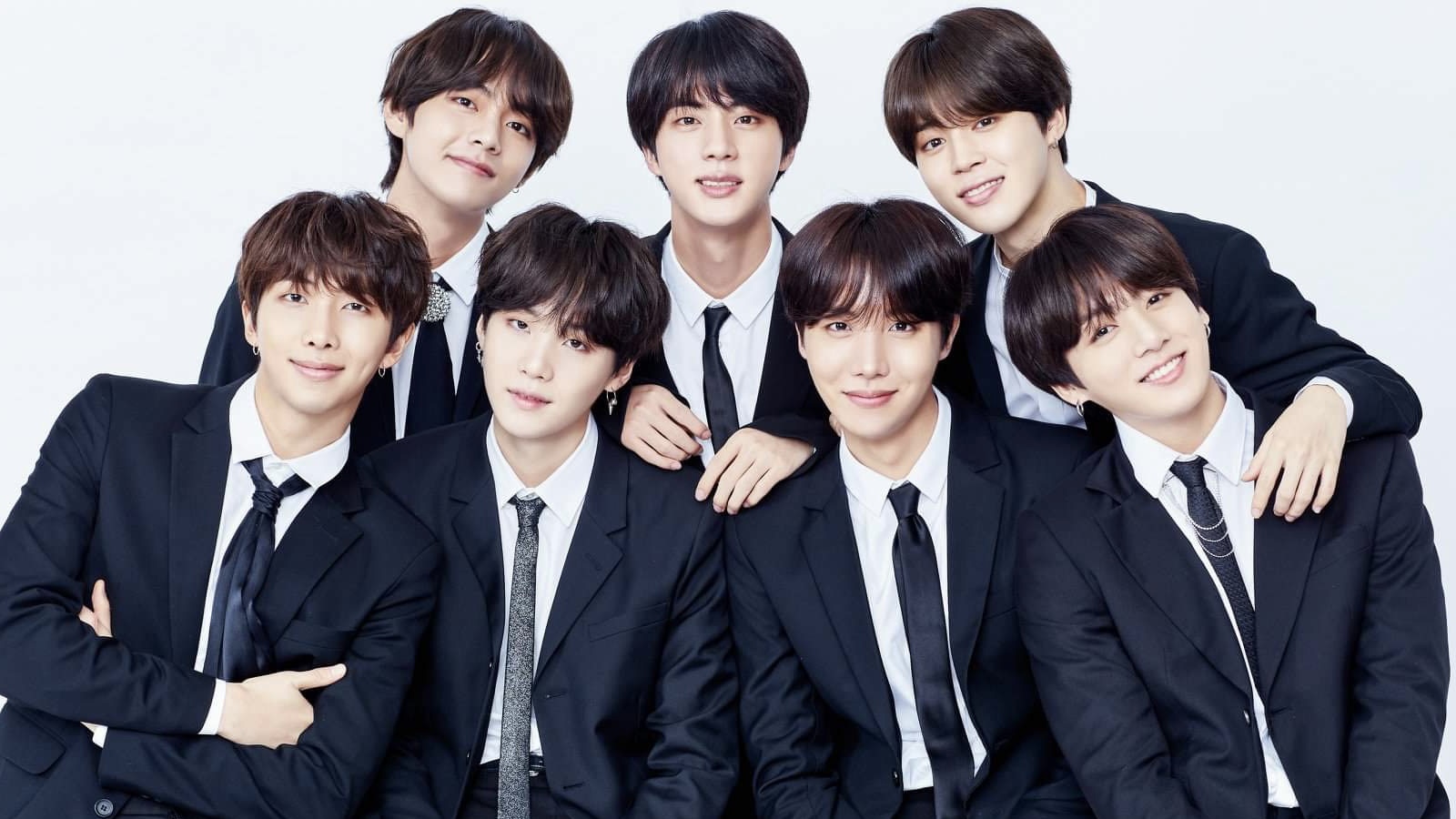 A foto apresenta todos os integrantes do BTS, grupo sul-coreano de k-pop, vestidos com paletó e gravata. Há três membros do grupo em pé e quatro à frente. A foto ilustra a matéria BTS: entenda caso de racismo que o grupo sofreu por radialista alemão"
