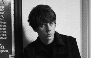A imagem apresenta o cantor Jake Bugg, que anunciou seu novo álbum "Saturday Night, Sunday Morning" na última sexta-feira, 19 de março. A foto está em preto e branco, o cantor está olhando para a frente e usando um casaco preto.
