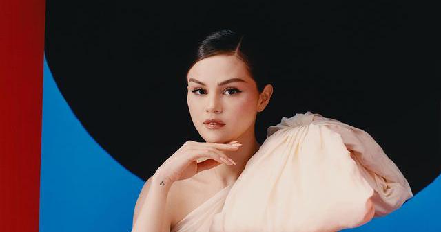 selena gomez divulgando seu álbum em espanhol. A cantora veste um vestido rosa claro, o cabelo preto amarrado, a mão queixo e atrás uma parede pintada com um círculo preto