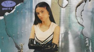 A imagem mostra um recorte da capa do single "good 4 u" da cantora Olivia Rodrigo. Ela está usando luvas pretas com os braços cruzados, usando uma roupa de líder de torcida e olhando para frente séria.