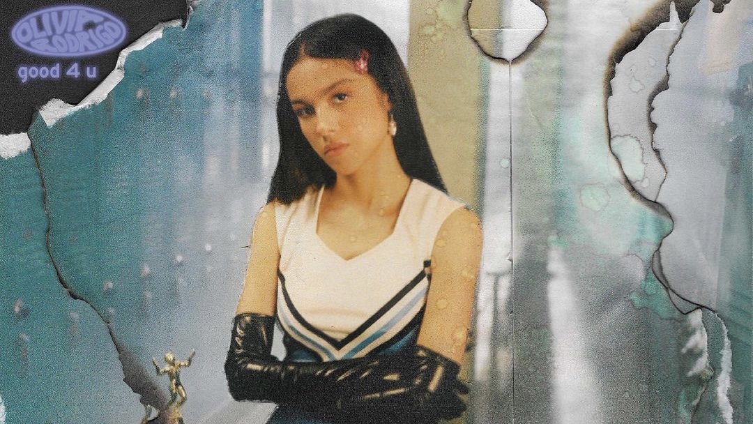 A imagem mostra um recorte da capa do single "good 4 u" da cantora Olivia Rodrigo. Ela está usando luvas pretas com os braços cruzados, usando uma roupa de líder de torcida e olhando para frente séria.
