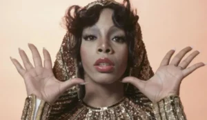 A imagem mostra Donna Summer com expressão marcante, vestindo uma roupa dourada brilhante com capuz, que lembra um estilo glamouroso e disco. Suas mãos estão erguidas próximas ao rosto, e ela usa maquiagem sofisticada, com destaque para os olhos e os lábios brilhantes. O fundo é de tom quente, criando um contraste elegante com o figurino cintilante.