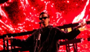 A imagem mostra Mark Foster durante sua apresentação no Lollapalooza Brasil 2025. Ele está no centro do palco, usando óculos escuros e uma jaqueta preta.