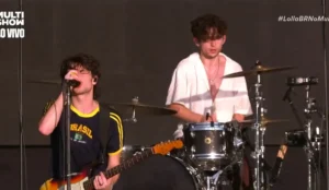 A imagem mostra um momento da apresentação da banda Inhaler no Lollapalooza Brasil 2025. No centro, o vocalista e guitarrista Elijah Hewson canta ao microfone enquanto toca guitarra. Ele veste uma camiseta retrô da seleção brasileira de futebol, demonstrando conexão com o público brasileiro. Ao fundo, o baterista Ryan McMahon toca sua bateria, vestindo uma camisa branca aberta. O palco tem uma iluminação simples, destacando os músicos. No canto superior esquerdo, há a marca do canal Multishow, que transmite o festival ao vivo, e no canto superior direito, a hashtag "#LollaBRNoMultishow".