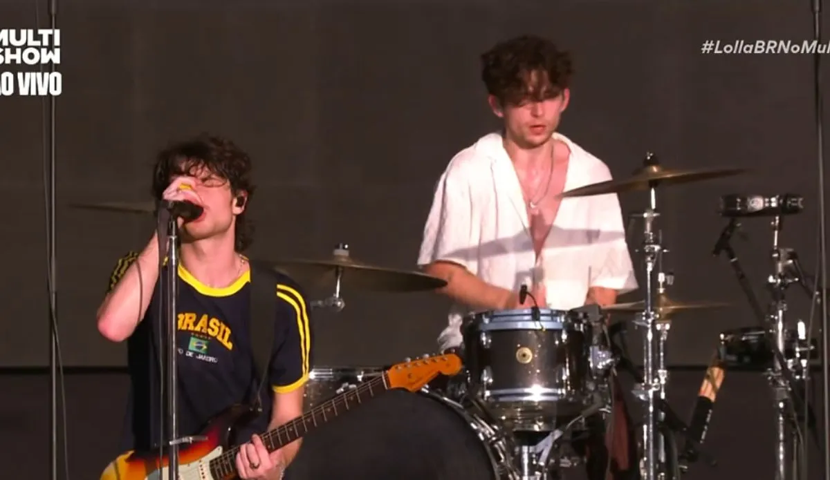 A imagem mostra um momento da apresentação da banda Inhaler no Lollapalooza Brasil 2025. No centro, o vocalista e guitarrista Elijah Hewson canta ao microfone enquanto toca guitarra. Ele veste uma camiseta retrô da seleção brasileira de futebol, demonstrando conexão com o público brasileiro. Ao fundo, o baterista Ryan McMahon toca sua bateria, vestindo uma camisa branca aberta. O palco tem uma iluminação simples, destacando os músicos. No canto superior esquerdo, há a marca do canal Multishow, que transmite o festival ao vivo, e no canto superior direito, a hashtag "#LollaBRNoMultishow".