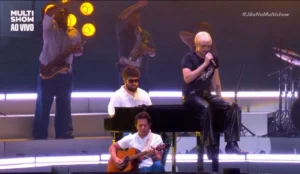 A imagem mostra um momento do show do cantor Jão no Lollapalooza Brasil 2025, transmitido ao vivo pelo Multishow. Jão, de cabelo loiro platinado, veste uma camiseta preta e calça de couro, e está sentado sobre um piano enquanto canta segurando um microfone. À sua frente, um músico toca violão, e outro, de óculos escuros e camisa branca, está ao piano. Ao fundo, há uma seção de metais com saxofonistas e trompetistas, e uma iluminação quente em tons de amarelo e laranja destaca a cena. A hashtag "#JãoNoMultishow" aparece no canto superior direito da imagem.