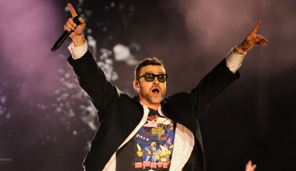 A imagem mostra o Justin Timberlake no palco durante uma apresentação ao vivo no Lollapalooza Brasil 2025. Ele veste uma jaqueta preta por cima de uma camisa listrada e uma camiseta estampada, além de usar óculos escuros. O artista segura um microfone em uma das mãos enquanto levanta os braços. O fundo da imagem tem uma iluminação vibrante, com tons escuros e efeitos de luz.
