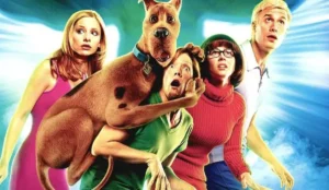 Imagem promocional do filme *Scooby-Doo* (2002), mostrando os personagens principais em uma cena de suspense. Scooby-Doo, o cachorro da turma, está assustado e agarrado em Salsicha, que também expressa medo. Atrás deles, Velma, Fred e Daphne olham para frente com expressões de surpresa e apreensão. O fundo apresenta uma luz azul brilhante.