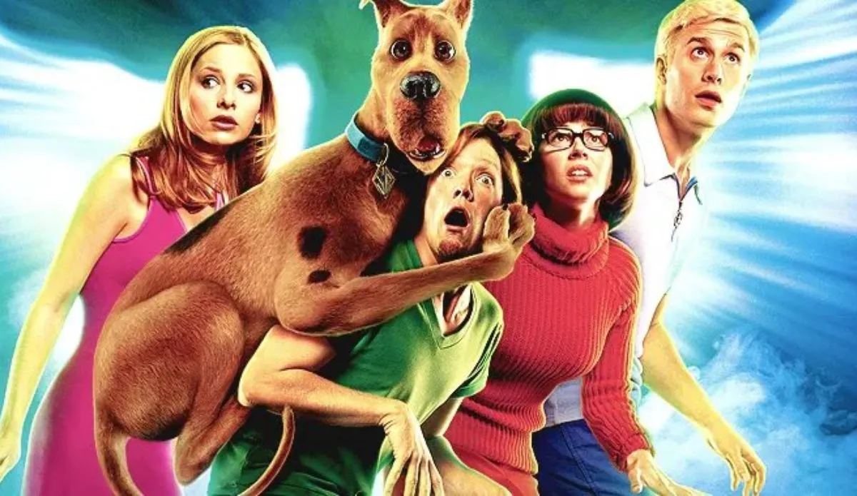 Imagem promocional do filme *Scooby-Doo* (2002), mostrando os personagens principais em uma cena de suspense. Scooby-Doo, o cachorro da turma, está assustado e agarrado em Salsicha, que também expressa medo. Atrás deles, Velma, Fred e Daphne olham para frente com expressões de surpresa e apreensão. O fundo apresenta uma luz azul brilhante.