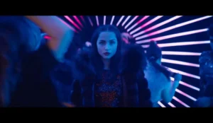 A imagem mostra uma cena do filme "Bailarina", spin-off da franquia "John Wick", estrelado por Ana de Armas. A atriz aparece no centro da imagem, vestindo um casaco de pele e um vestido brilhante, com uma expressão séria e determinada. O fundo exibe uma ambientação de boate, com luzes neon dispostas em formato radial, criando um efeito visual dinâmico. Outras pessoas estão dançando ao redor, mas desfocadas, dando destaque à personagem de Ana de Armas. A iluminação azul e rosa realça o clima noturno e estilizado da cena, remetendo ao tom visual da franquia "John Wick".