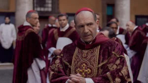Cena do filme "Conclave" com Ralph Fiennes caracterizado como cardeal, cercado por outros religiosos vestindo trajes litúrgicos