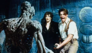 Cena do filme “A Múmia” (1999), com Brendan Fraser gritando enquanto aponta uma arma para a criatura ameaçadora à sua frente; ao seu lado, Rachel Weisz observa a cena assustada, em meio a um cenário com inscrições egípcias antigas