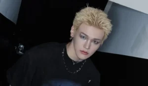 Bain, integrante do grupo de K-pop Just B, aparece na imagem com cabelo loiro curto e espetado. Ele veste uma camisa preta e um colar de corrente. O fundo é escuro, com algumas áreas iluminadas, e Bain está posicionado diante de uma porta com uma tranca visível