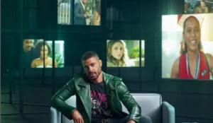 Cauã Reymond está sentado em um sofá cinza, vestindo uma jaqueta de couro verde e uma camiseta gráfica preta. Ao fundo, há várias telas exibindo imagens desfocadas de personagens da novela "Vale Tudo"