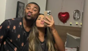 Davi Brito e sua namorada Adriana Paula posam juntos em frente ao espelho, fazendo biquinho para a câmera; ao fundo, há balões decorativos em formato de coração e de letra