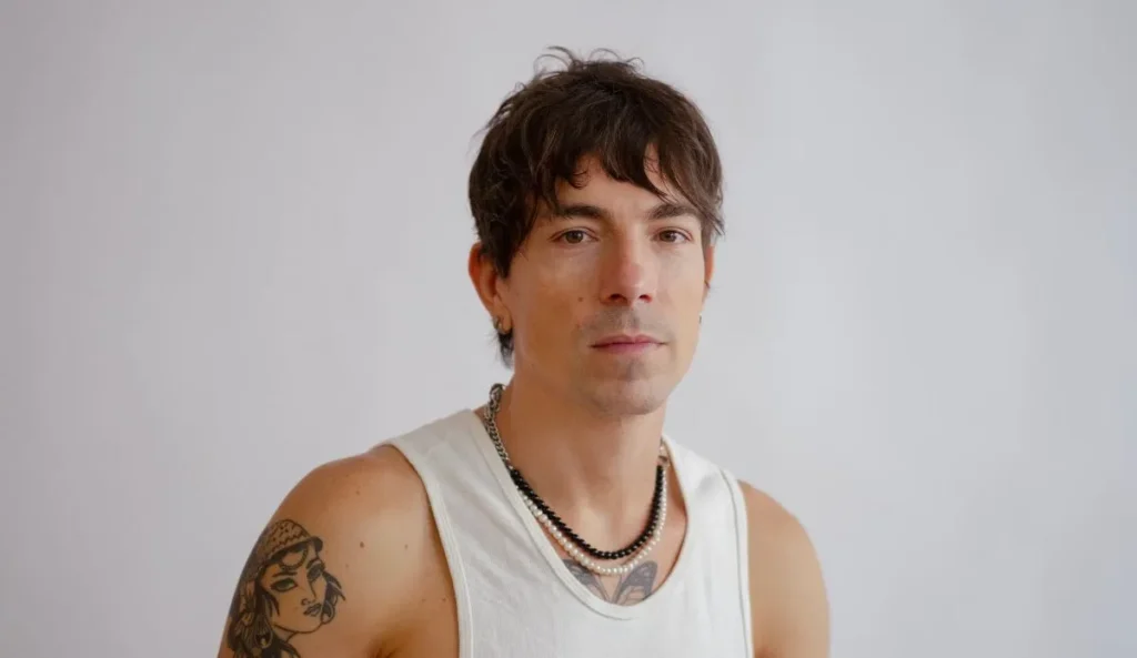 Di Ferrero com expressão séria, vestindo regata branca e colares em camadas. O cantor está posando diante de um fundo neutro, com tatuagens visíveis no braço esquerdo