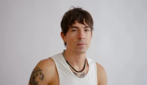 Di Ferrero com expressão séria, vestindo regata branca e colares em camadas. O cantor está posando diante de um fundo neutro, com tatuagens visíveis no braço esquerdo