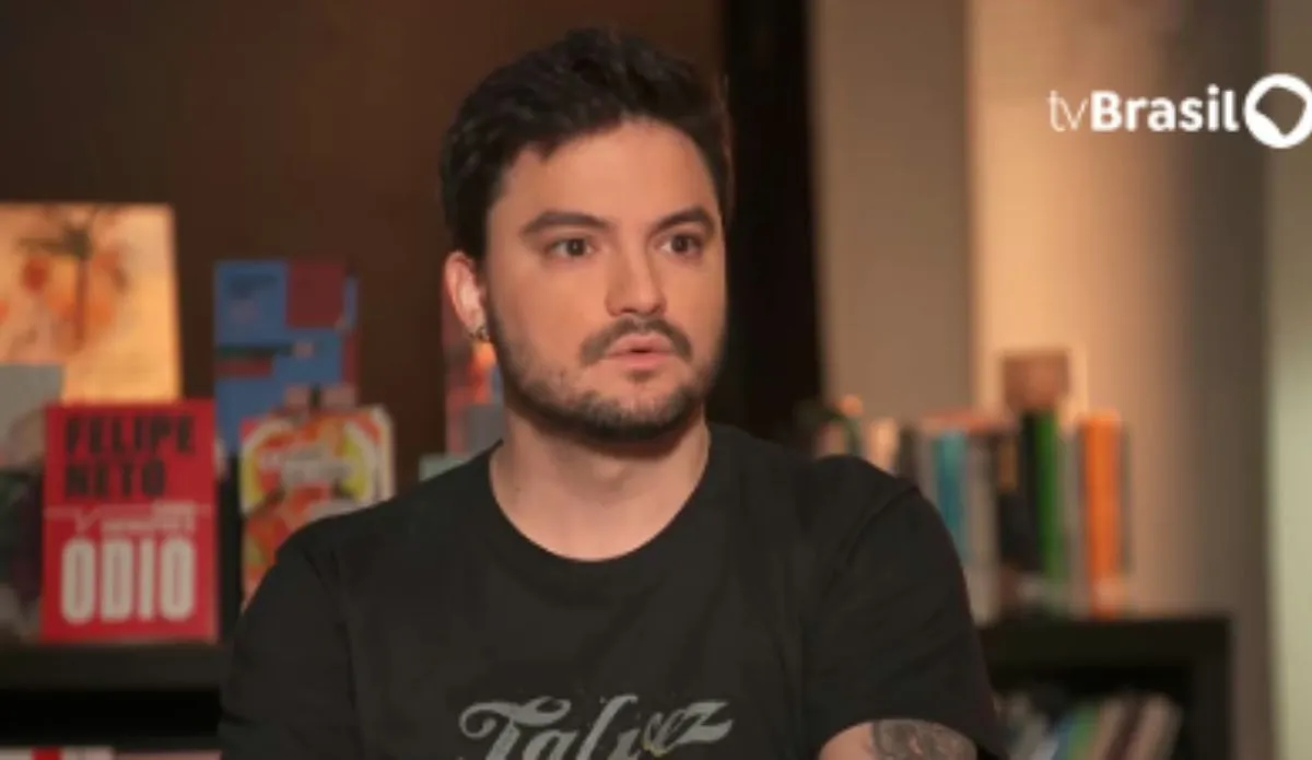 Captura de tela de uma entrevista com Felipe Neto na TV Brasil. O influenciador aparece de barba curta, cabelo escuro e veste uma camiseta preta com a palavra "Talvez" estampada. Ao fundo, há uma estante com livros, incluindo um de sua autoria intitulado "Como Enfrentar o Ódio"