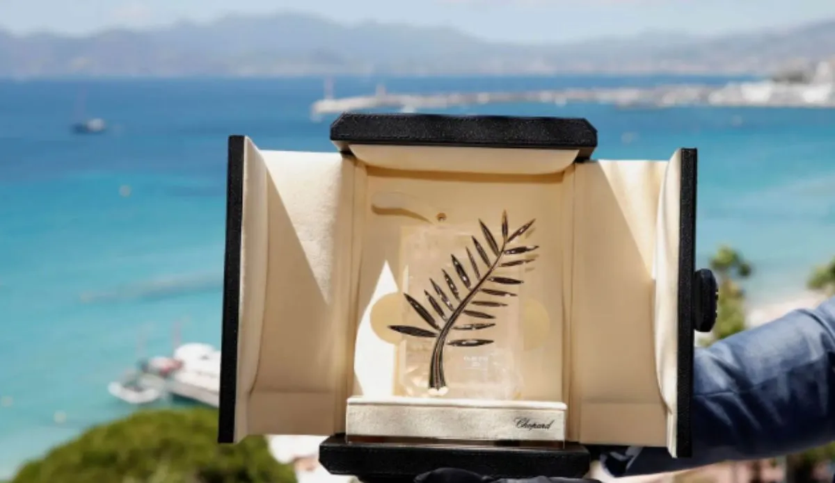 Troféu da Palma de Ouro do Festival de Cannes, exibido em uma caixa aberta com fundo do mar azul e costa da cidade ao fundo