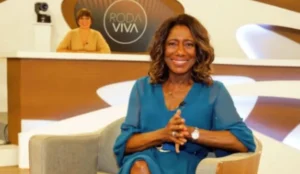 Gloria Maria sorrindo enquanto participa do programa Roda Viva; ela está sentada em uma poltrona com as mãos cruzadas, vestindo um vestido azul, com o logo do programa ao fundo