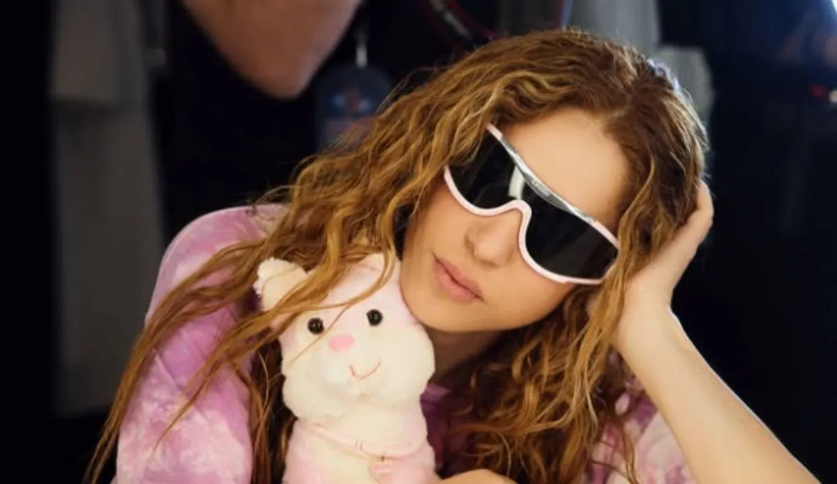 Na imagem, Shakira aparece com um visual descontraído e estiloso. A cantora usa óculos escuros com armação branca e lentes espelhadas, combinando com seu look em tons de rosa e lilás. Seu cabelo cacheado solto reforça sua estética natural e marcante. Ela segura um ursinho de pelúcia rosa com um colar brilhante