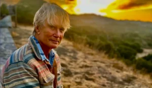 Richard Norton sentado ao ar livre durante o pôr do sol, vestindo uma jaqueta listrada e uma camisa azul, com uma paisagem cênica de colinas e árvores ao fundo, sob um céu vibrante em tons de amarelo e laranja