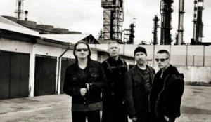Integrantes da banda U2 posam juntos em área industrial, com prédios e chaminés ao fundo; todos usam roupas escuras e encaram a câmera com expressão séria