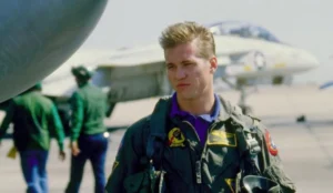 A imagem mostra Val Kilmer no set de Top Gun, vestido como piloto de caça com um uniforme cheio de patches e insígnias, incluindo um com a inscrição "TOP GUN". Ao fundo, há um avião de caça.