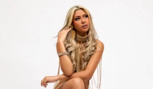 A cantora Wenny posa para a foto com cabelo loiro comprido e ondulado, usa um conjunto glamoroso de correntes douradas e acessórios, criando uma aparência luxuosa. Ela posa com uma mão apoiada no braço oposto, que está dobrado. O fundo branco realça o foco na elegância e estilo de Wenny