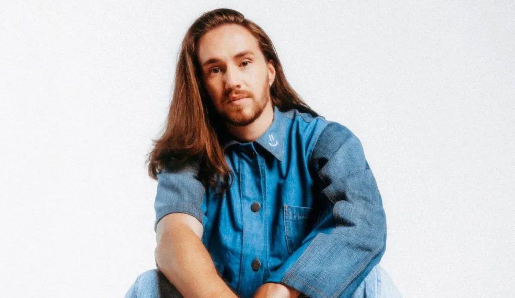 Vitor Kley, em foto para divulgação do álbum "As Pequenas Grandes Coisas", sentado em um fundo branco, vestindo uma jaqueta jeans azul. Ele está com os braços apoiados nos joelhos e olha diretamente para a câmera, com os cabelos longos soltos.