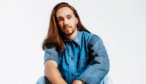 Vitor Kley, em foto para divulgação do álbum "As Pequenas Grandes Coisas", sentado em um fundo branco, vestindo uma jaqueta jeans azul. Ele está com os braços apoiados nos joelhos e olha diretamente para a câmera, com os cabelos longos soltos.