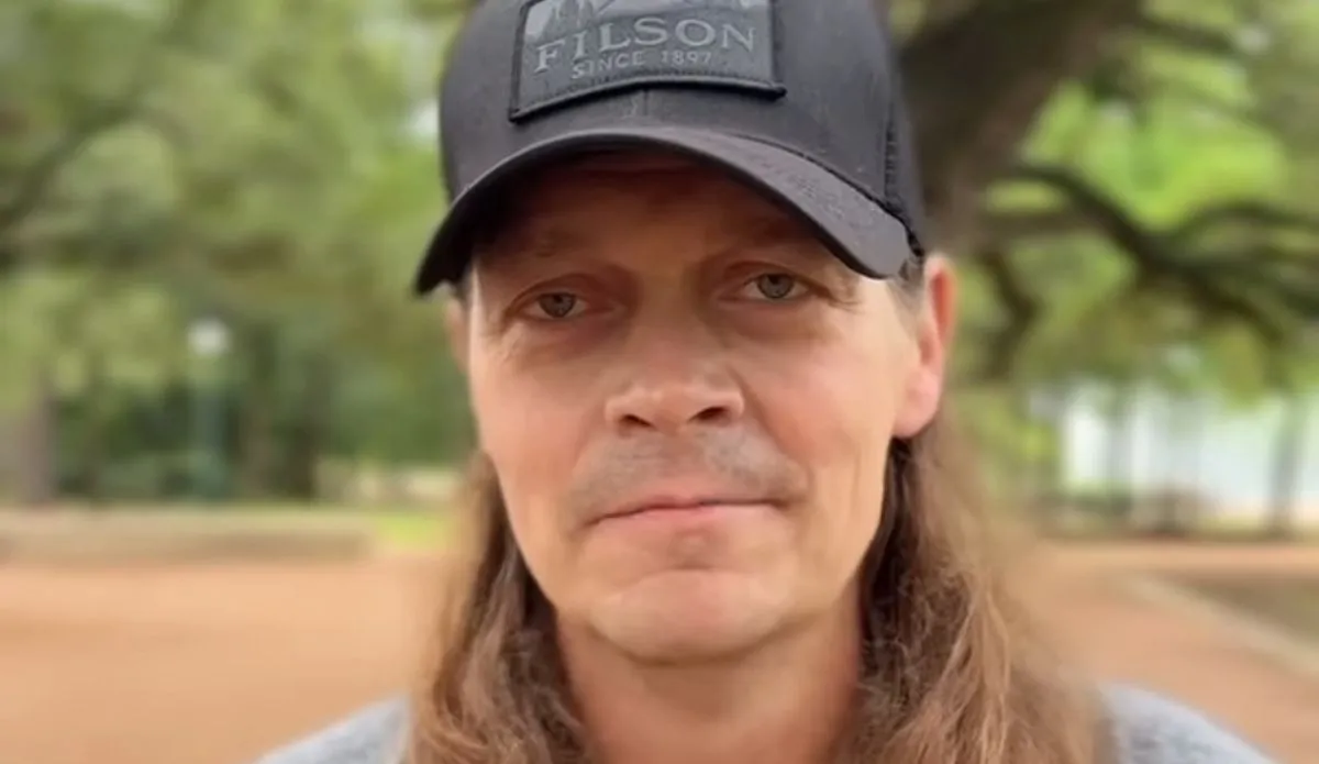 Brad Arnold, vocalista da banda 3 Doors Down, usando boné preto e olhando para a câmera, em meio a árvores ao fundo.