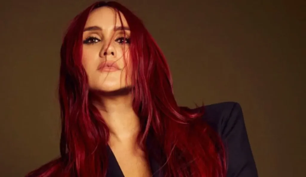 A cantora Dulce María, de cabelos ruivos vibrantes e olhar expressivo, vestindo um blazer escuro contra um fundo neutro.