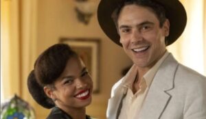 Sergio Guizé e Jeniffer Nascimento em cena da novela “Êta Mundo Melhor!”, da TV Globo. Os personagens aparecem sorridentes, vestidos com figurinos de época, em ambiente interno iluminado.