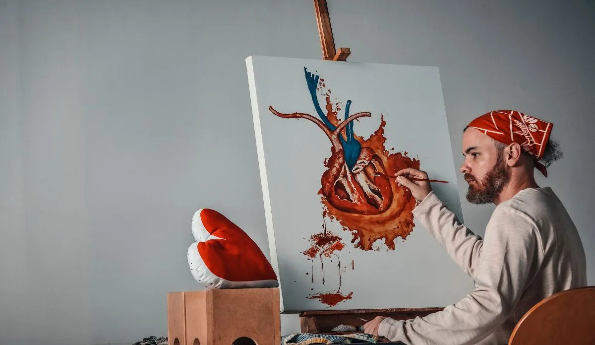 O artista Flavio Delli pinta uma obra que representa um coração anatômico com detalhes em vermelho e azul. Ele aparece concentrado, usando uma bandana vermelha, em frente a uma tela apoiada em cavalete, com um travesseiro em forma de coração ao lado.