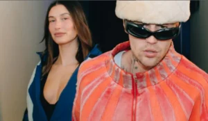 Justin Bieber e Hailey Bieber posam juntos; ele veste casaco vermelho com óculos escuros e gorro, enquanto ela aparece ao fundo com jaqueta azul e expressão neutra.
