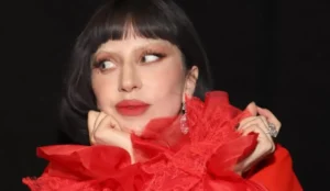 Lady Gaga usando um vestido vermelho volumoso com detalhes de tule, olhando para o lado com expressão curiosa; artista está confirmada no Tudum 2025